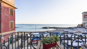 A balcony with a view of the sea, a red planter with green plants, and boats covered with tarps.