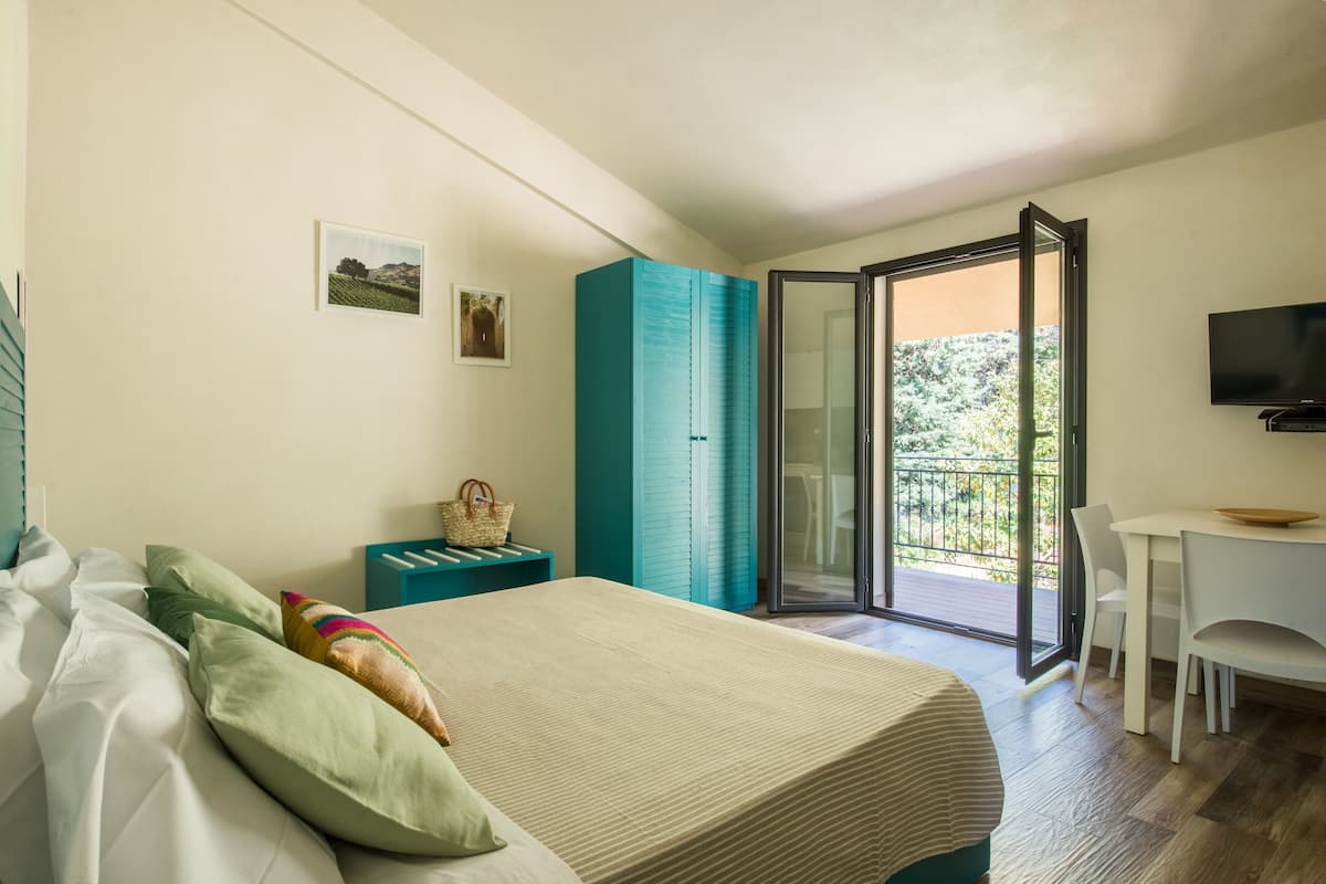 A bedroom with a bed, a desk, and a balcony with a view of greenery.
