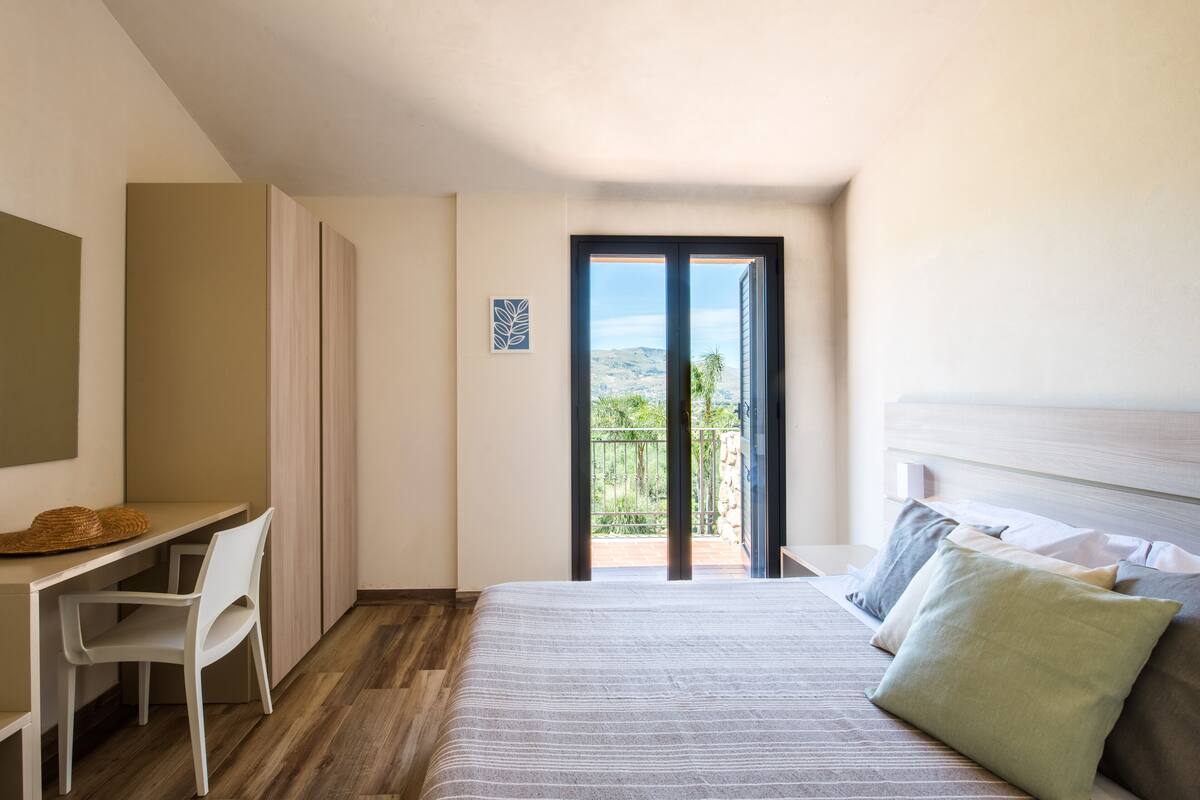 A bedroom with a bed, a desk, and a chair. There is a balcony with a view of the outdoors.