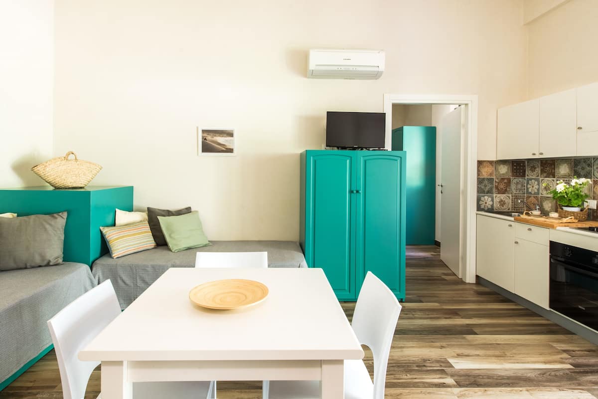 A compact living space with a teal sofa, a white dining table, and a kitchen area with teal cabinets and a tiled backsplash.