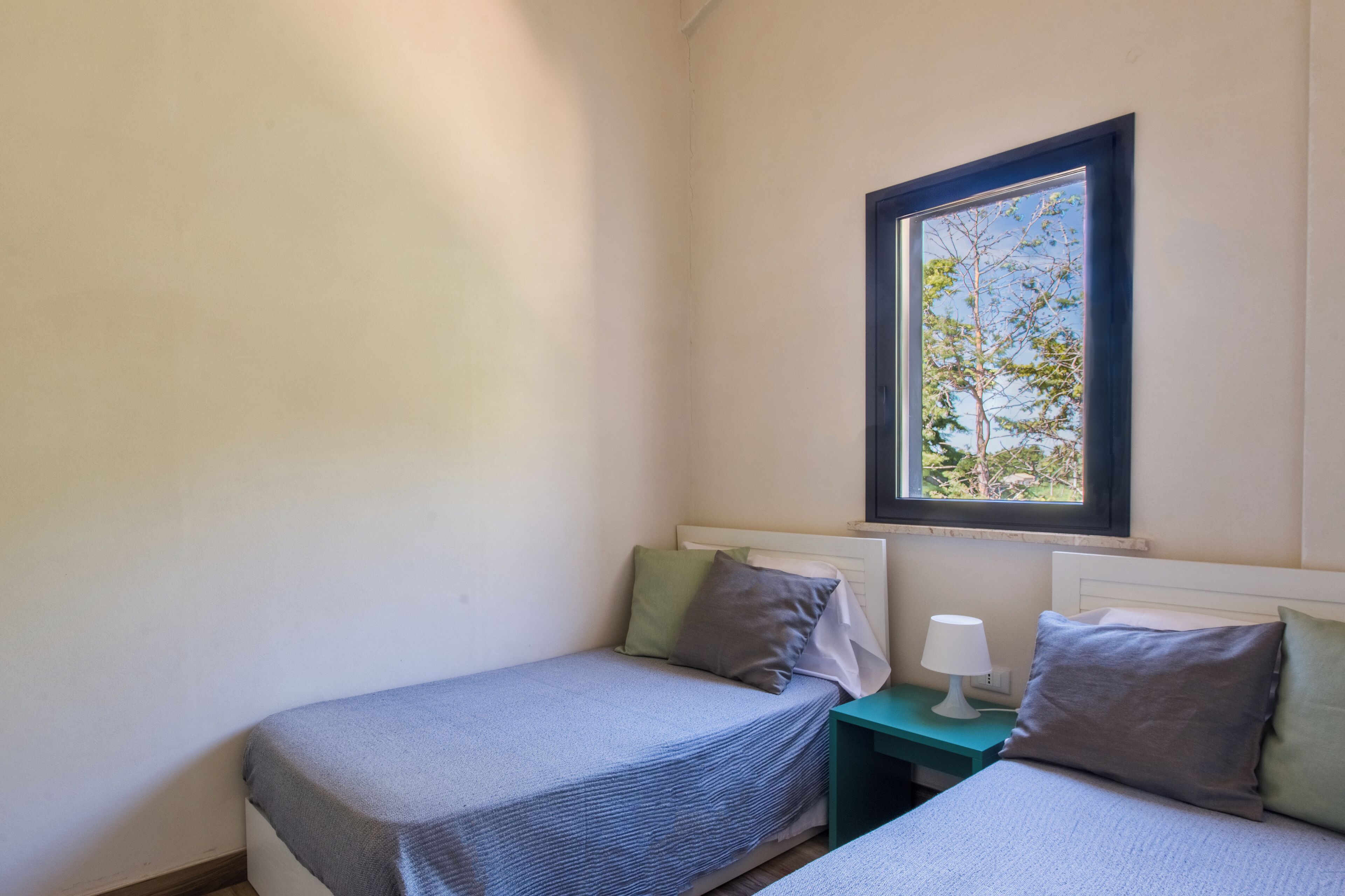 A small, clean room with two single beds, a nightstand with a lamp, and a window with a view of trees.
