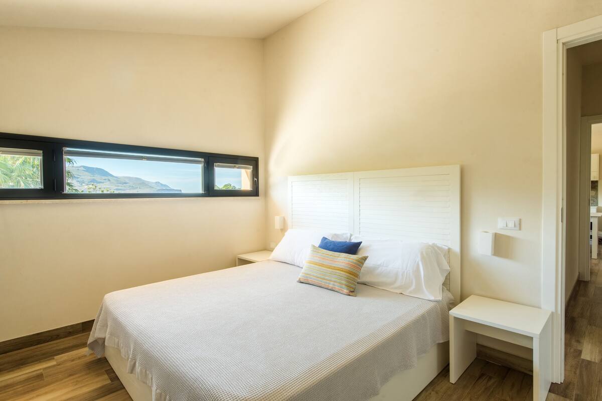 A bedroom with a bed, a nightstand, a window with a view, and a door.