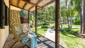 A covered outdoor seating area with stone walls, a thatched roof, and a view of a lush garden and hills.