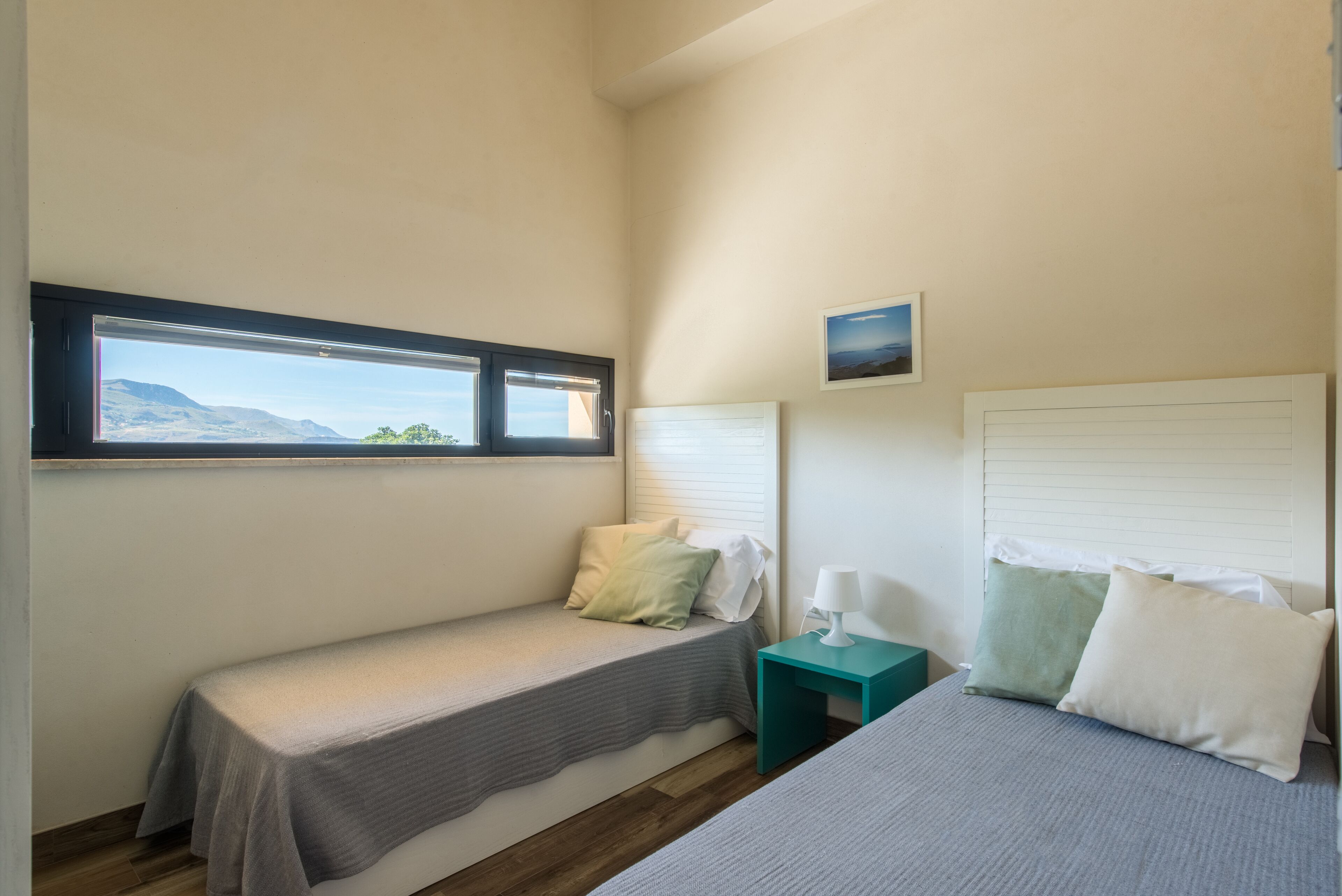 A bedroom with a bed, a small bedside table, a window with a view of mountains, and a wall-mounted picture.