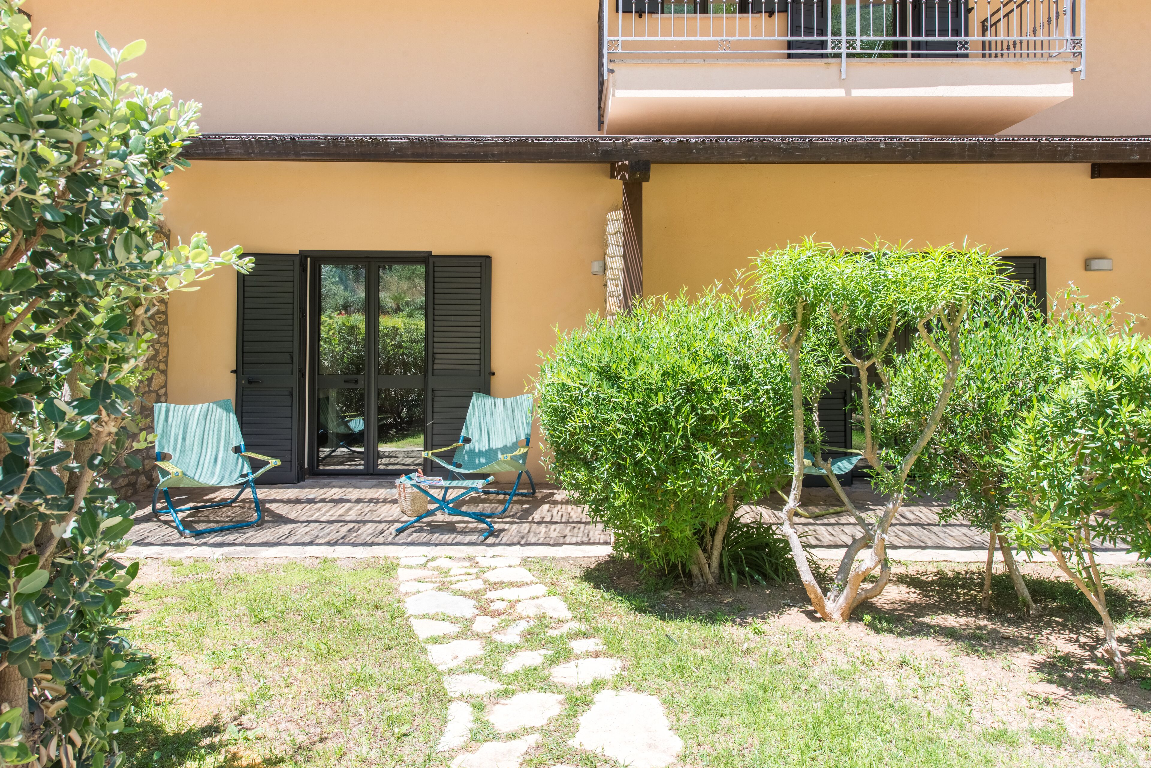 A building with a balcony, a door with shutters, and a stone pathway leading to a seating area with chairs.