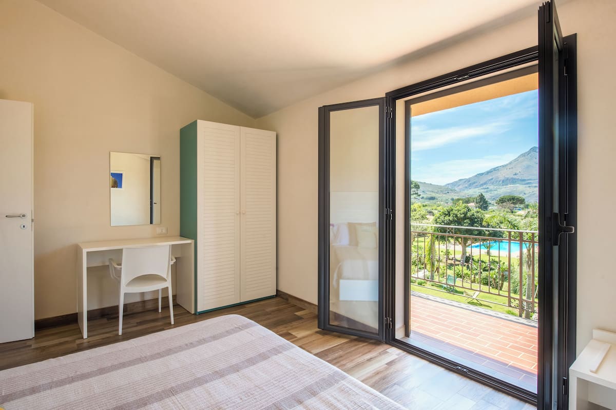 A bedroom with a bed, a desk, a chair, a wardrobe, and a sliding glass door leading to a balcony with a pool view.