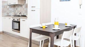 A modern kitchen with white cabinets, a stove, and a sink. There is a dining table with white chairs and a dark tablecloth. A painting is on the wall above the table.