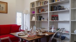 A dining area with a wooden table set for four, surrounded by clear chairs. A red sofa is against the wall, and a white bookshelf filled with books and decorative items is to the right. - Cozy Family Apartment in Castelletto by Wonderful Italy (Genoa)