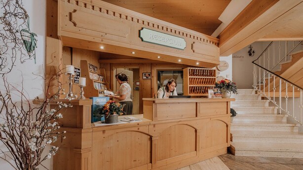 A wooden reception desk with a sign reading "Reception," a staircase with a railing, and a vase with flowers on the desk. - Ferienwohnung (Marquartstein)