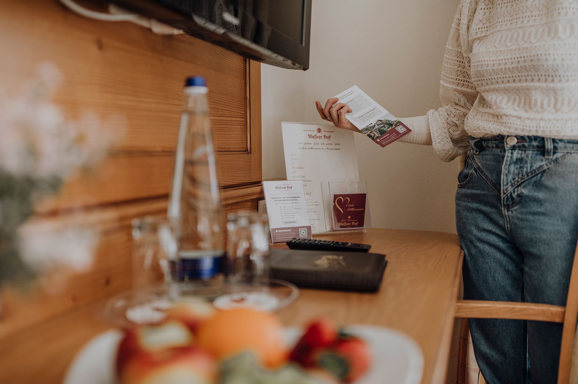 Une personne tenant une carte sur une table en bois, avec une bouteille, des verres et une assiette de fruits.