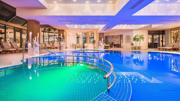 Una piscina interior con suelo de baldosas azules y borde curvo. Se observan sillones de descanso y una zona de estar con mesas y sillas.