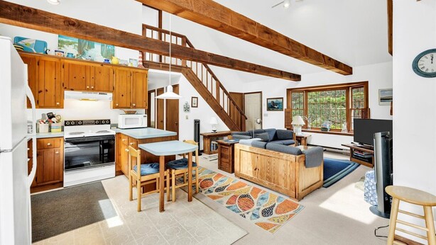 Une cuisine spacieuse avec des meubles en bois, un coin repas comprenant une table et des chaises, un salon confortable avec un canapé et une télévision, et un escalier menant à l’étage supérieur.