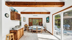 Une pièce chaleureuse avec un plafond en bois, un coin repas avec une table et des chaises, un coin cuisine avec un meuble à vin, et une grande porte-fenêtre coulissante donnant sur une terrasse extérieure.