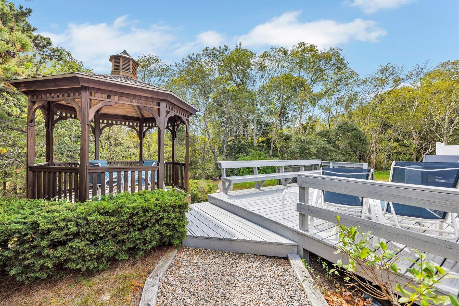 A wooden gazebo with a bench, surrounded by greenery and a wooden deck leading to a covered area.