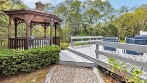 Un belvédère en bois avec un banc, entouré de verdure et doté d’une terrasse en bois menant à une zone abritée.