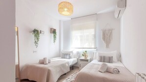 A minimalist bedroom with two beds, a bedside table, a hanging plant, and a large window. - Syros Apartment (Javea / Xàbia)