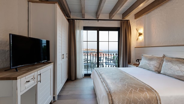 Habitación de hotel con cama, televisión sobre un mueble de madera, balcón con vistas y una lámpara de pared.