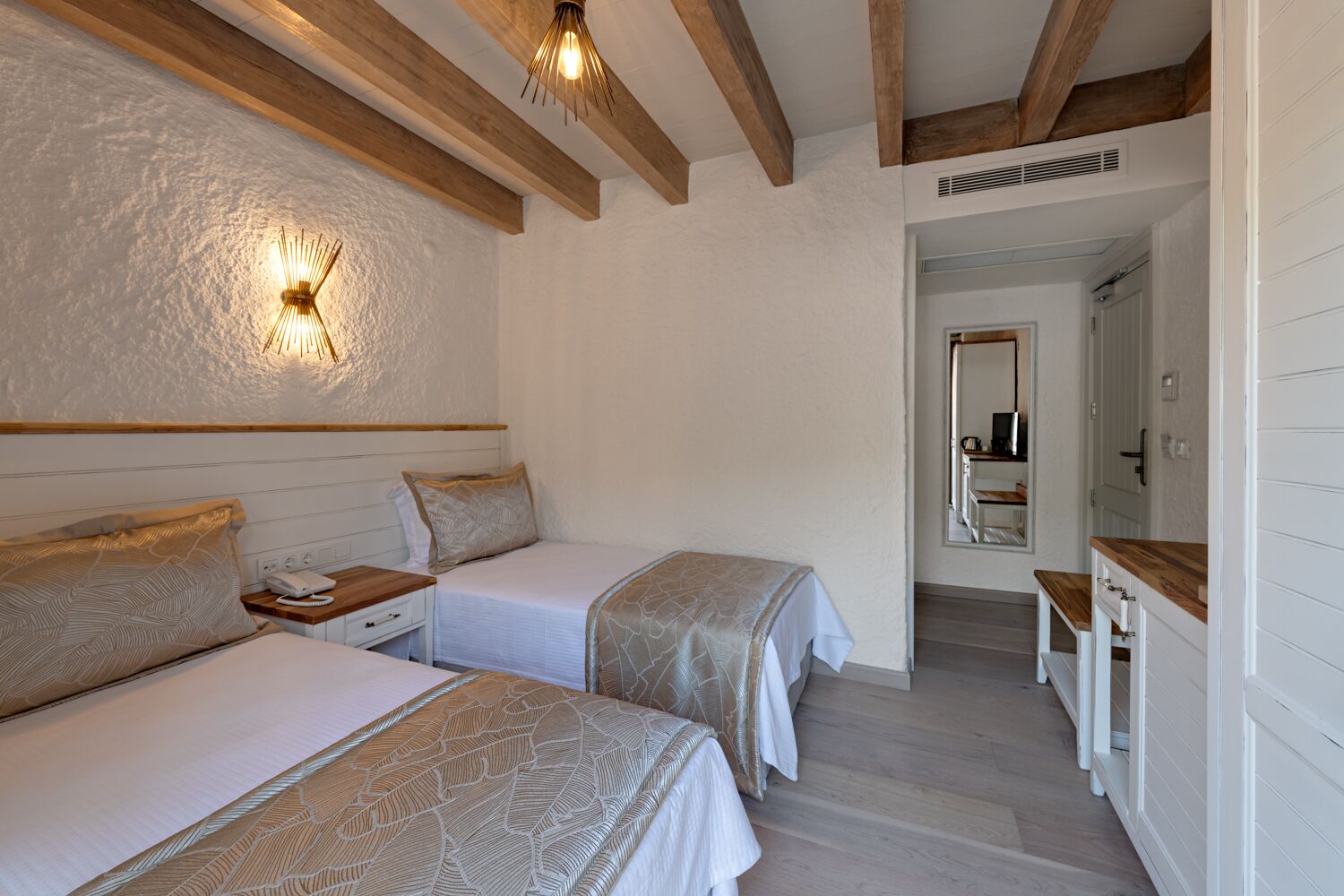 A bedroom with two beds, wooden beams, a wall-mounted light fixture, and a hallway leading to a bathroom.
