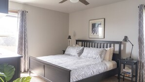 Une chambre à coucher comprenant un lit, des tables de chevet, un ventilateur de plafond, une fenêtre avec des rideaux et un tableau encadré accroché au mur.