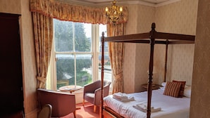 A four-poster bed with a canopy, a chaise lounge, a small round table, and a chair in a room with patterned wallpaper and large windows. - Portland Hotel (Buxton)