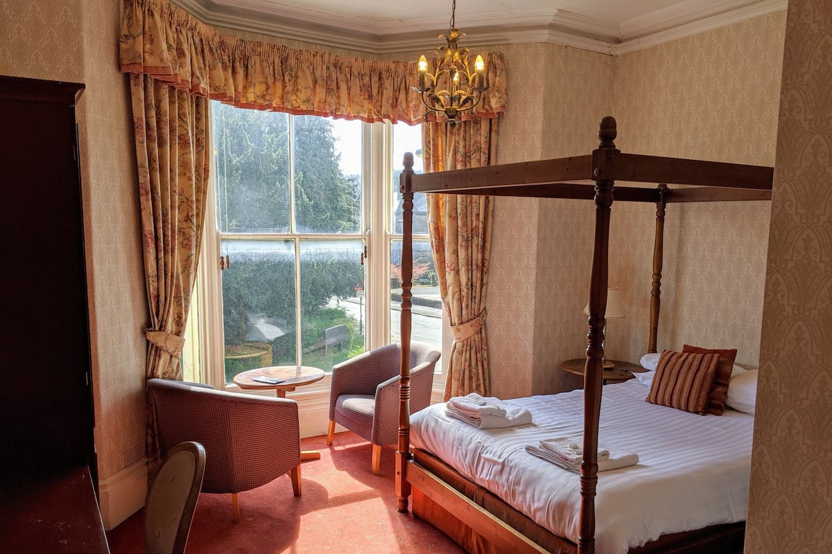 A four-poster bed with a canopy, a chaise lounge, a small round table, and a chair in a room with patterned wallpaper and large windows.