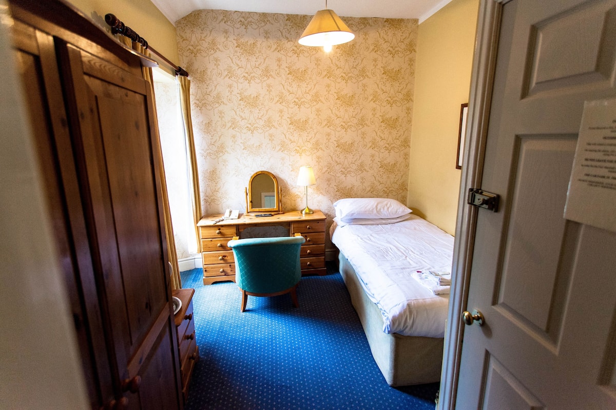A small, compact bedroom with a single bed, a desk with a mirror, and a chair.