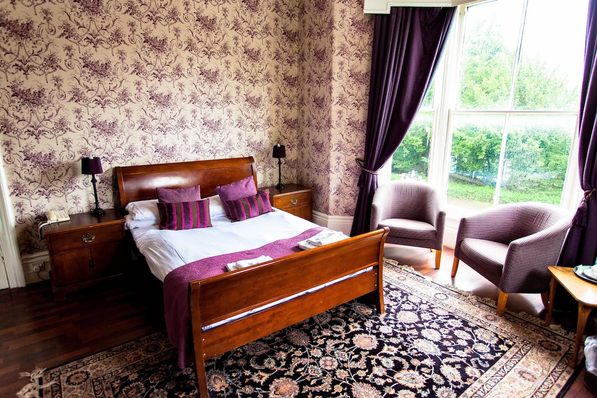A bedroom with a wooden bed, a nightstand, a chair, a window with curtains, and a patterned carpet.