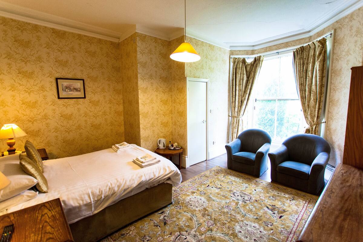 A hotel room with a bed, two armchairs, a small table with a lamp, a picture on the wall, and a window with curtains.