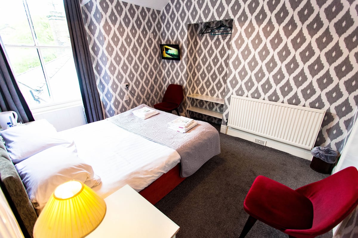 A hotel room with a bed, a red chair, a TV on the wall, and a window with curtains.