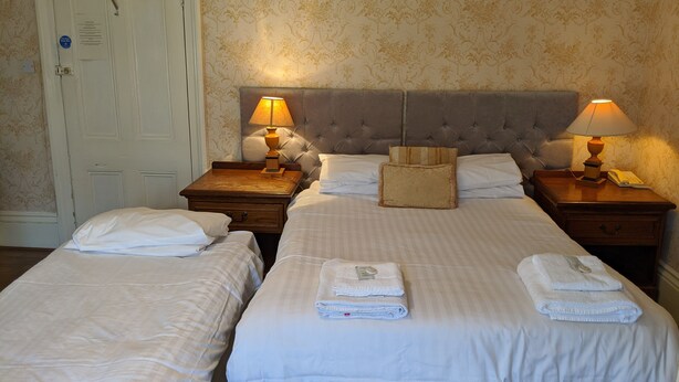 A hotel room with two single beds, a large bed, and a nightstand with a lamp. There is a door with a sign on it. - Portland Hotel (Buxton)
