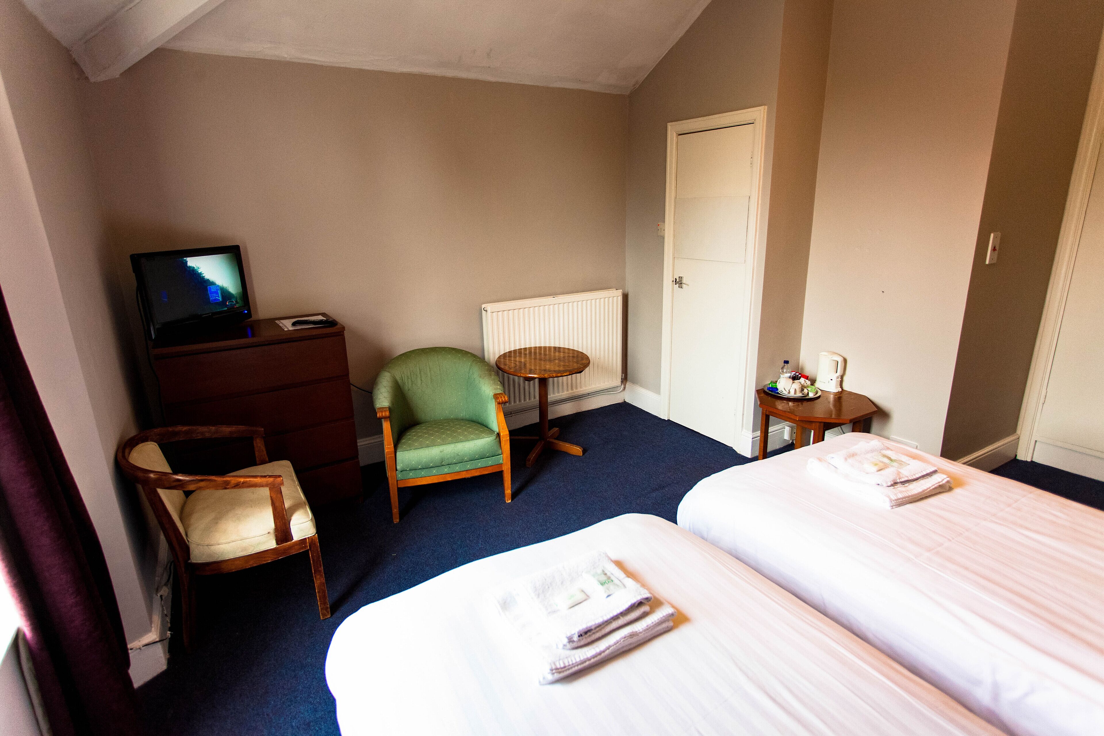 A hotel room with two beds, a television, a green armchair, a small round table, and a radiator.