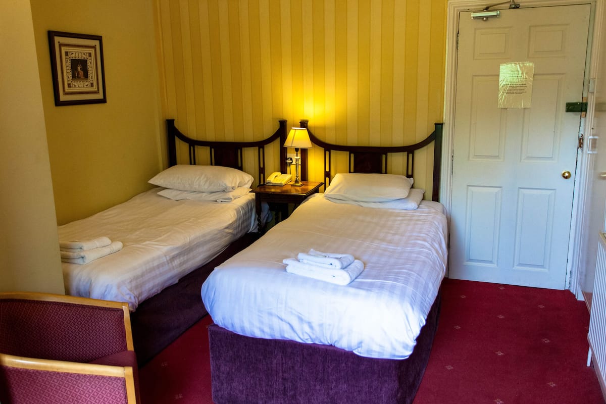 A hotel room with two single beds, a nightstand with a lamp, a framed picture on the wall, and a door with a notice.