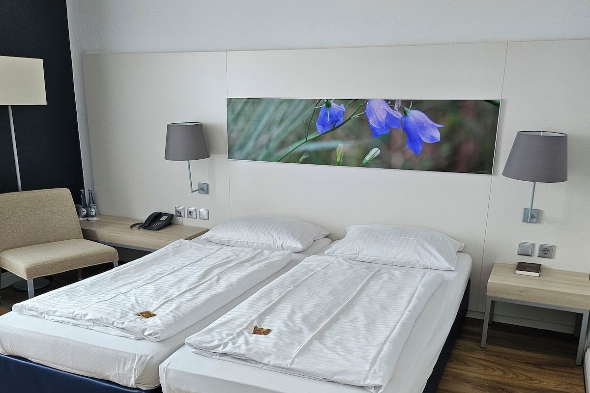 A double bed with white linens, two bedside tables with lamps, a wall-mounted phone, and a large framed picture of flowers.