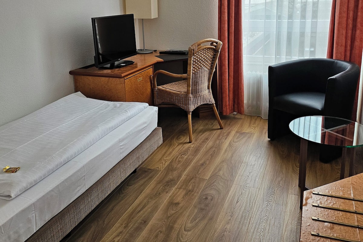 A hotel room with a single bed, a desk with a computer, a chair, a small table, and a view of the city through sheer curtains.