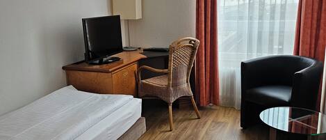 A hotel room with a single bed, a desk with a computer, a chair, a small table, and a view of the city through sheer curtains.