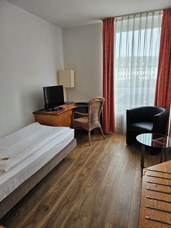 Habitación de hotel con una cama individual, un escritorio con ordenador, una silla, una mesita y vistas a la ciudad a través de cortinas translúcidas.