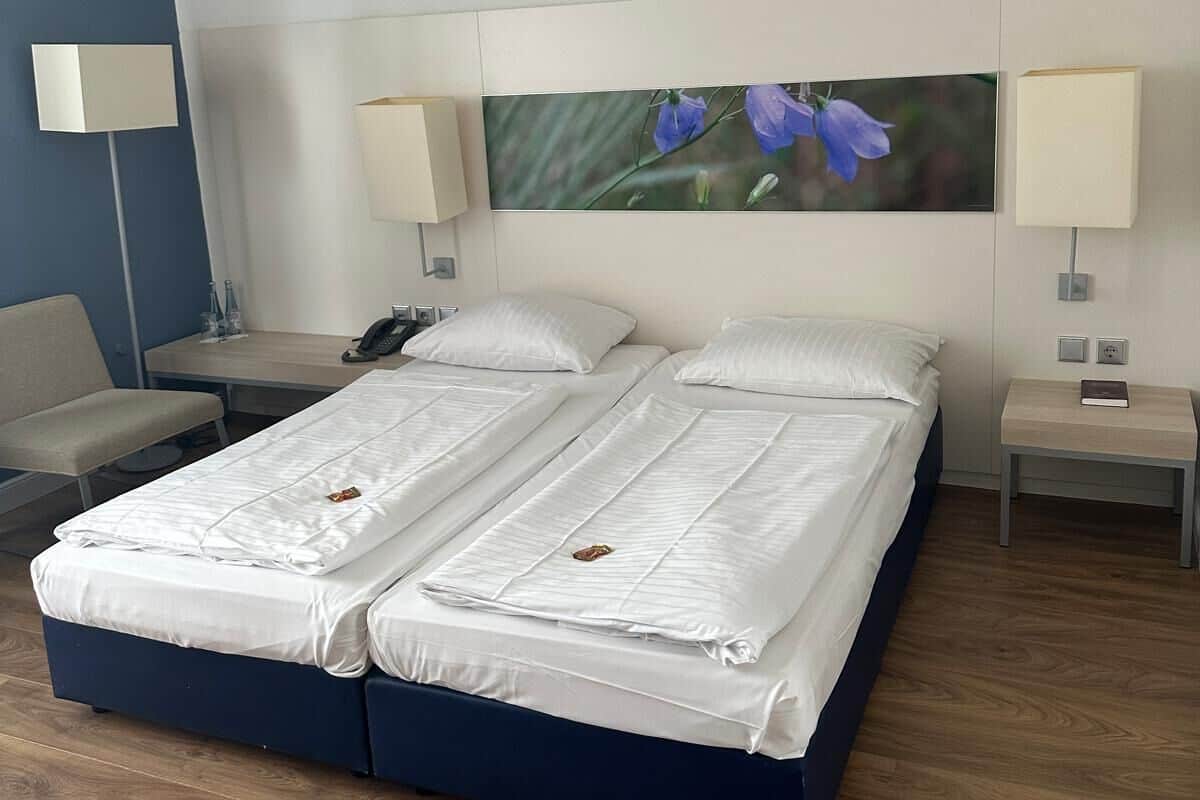 A double bed with white linens, two bedside tables with lamps, a chair, and a wall-mounted picture of flowers.