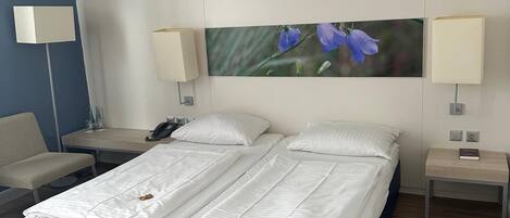 Cama doble con ropa blanca, dos mesitas de noche con lámparas, una silla y un cuadro de flores colgado en la pared.