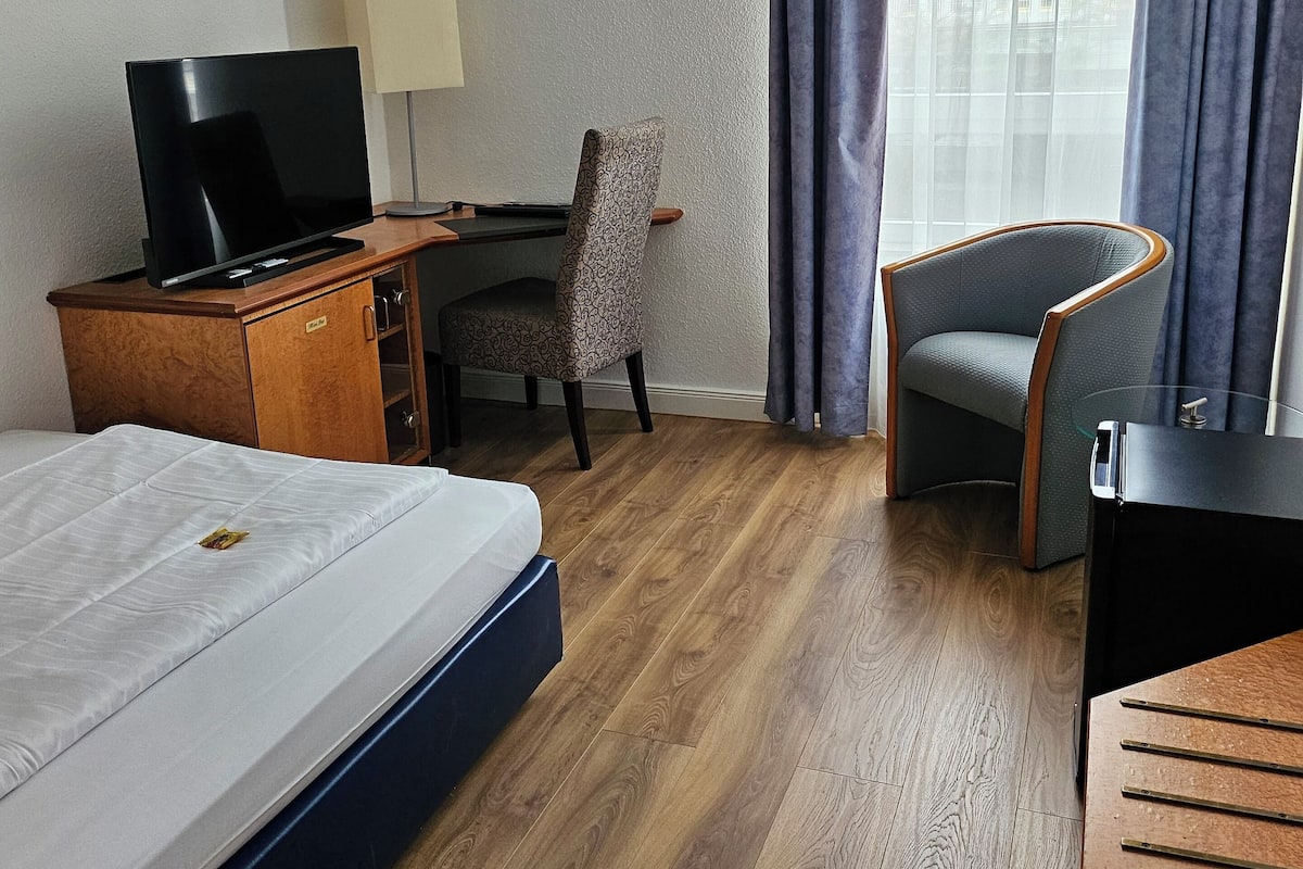 A hotel room with a bed, a desk with a TV, a chair, and a desk lamp.
