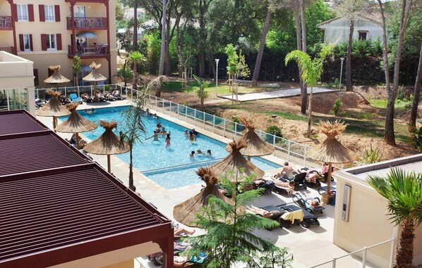 A swimming pool with lounge chairs and umbrellas, surrounded by trees and a building with balconies. - Cozy & Contemporary Stay (Cavalaire-sur-Mer)