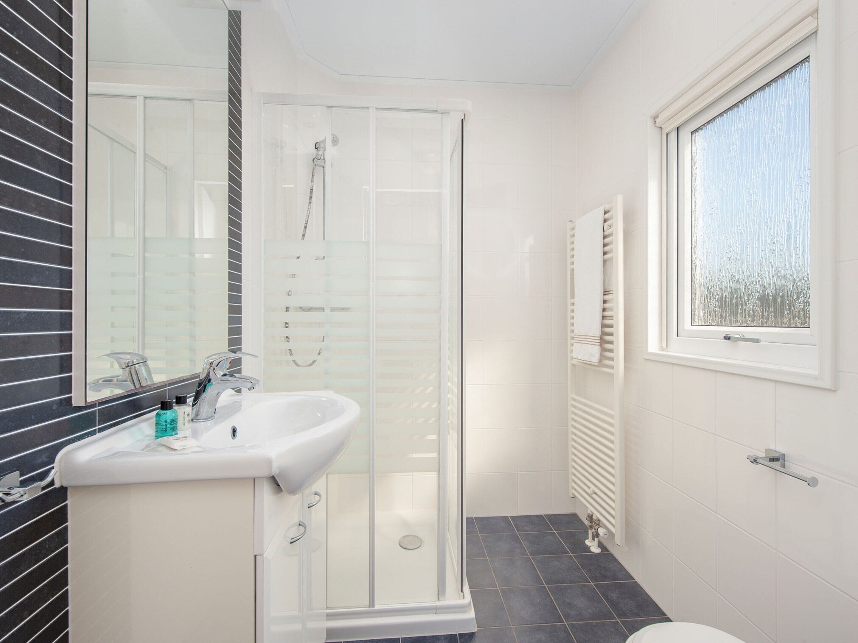 A modern bathroom with a glass shower enclosure, a white sink, and a window with blinds.