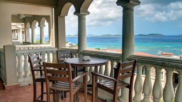 A balcony with a table and chairs, overlooking a coastal view with clear skies and distant islands.