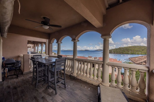 A balcony with a table and chairs, a ceiling fan, and a view of the sea and buildings.