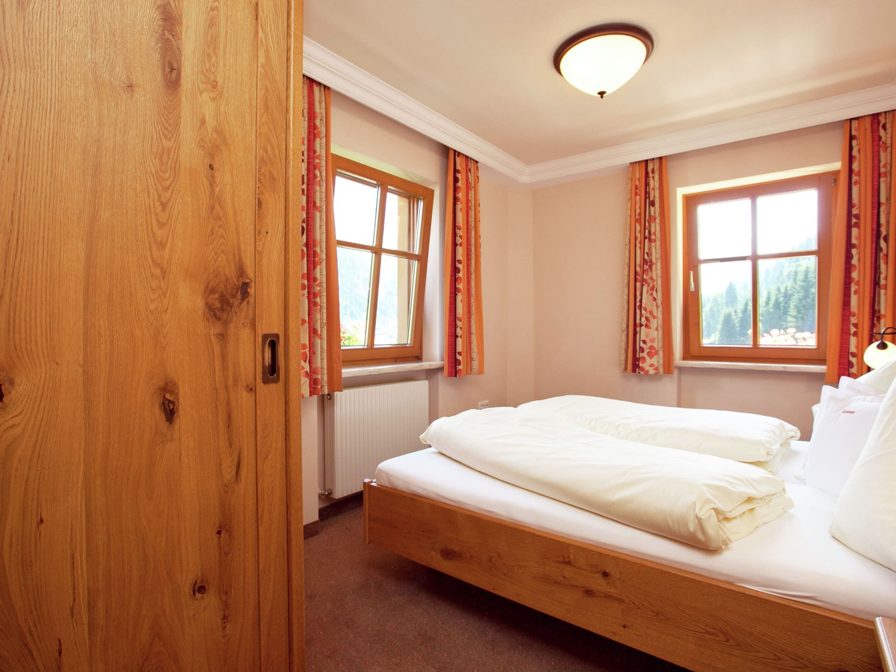Une chambre à coucher avec un lit en bois, deux fenêtres avec des rideaux, un radiateur et un luminaire au plafond.