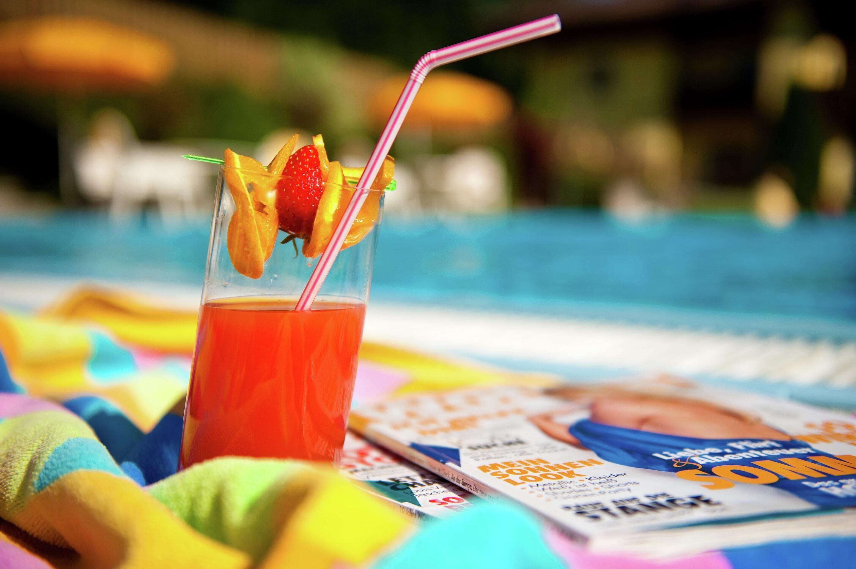 A glass of red beverage with fruit garnish and a straw, placed on a colorful towel next to a magazine.