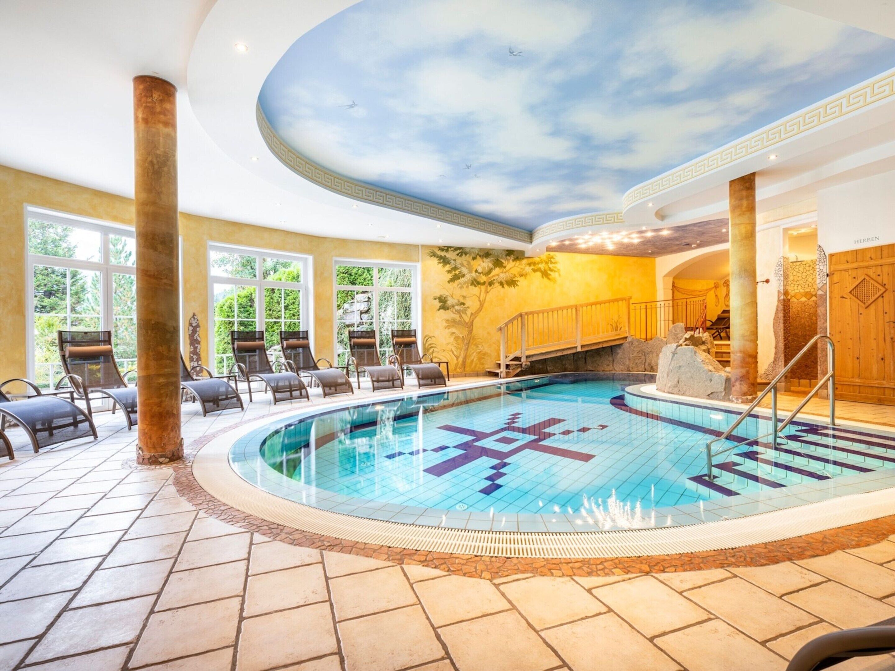 An indoor swimming pool with a ceiling mural, sun loungers, and a stone-tiled floor.