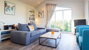 A modern living room with a grey sofa, a wooden coffee table, and a blue armchair. There is a bookshelf with books and a flat-screen TV on a stand. - Neat Chalet With Microwave Near the Beach in Egmond (Egmond aan den Hoef)