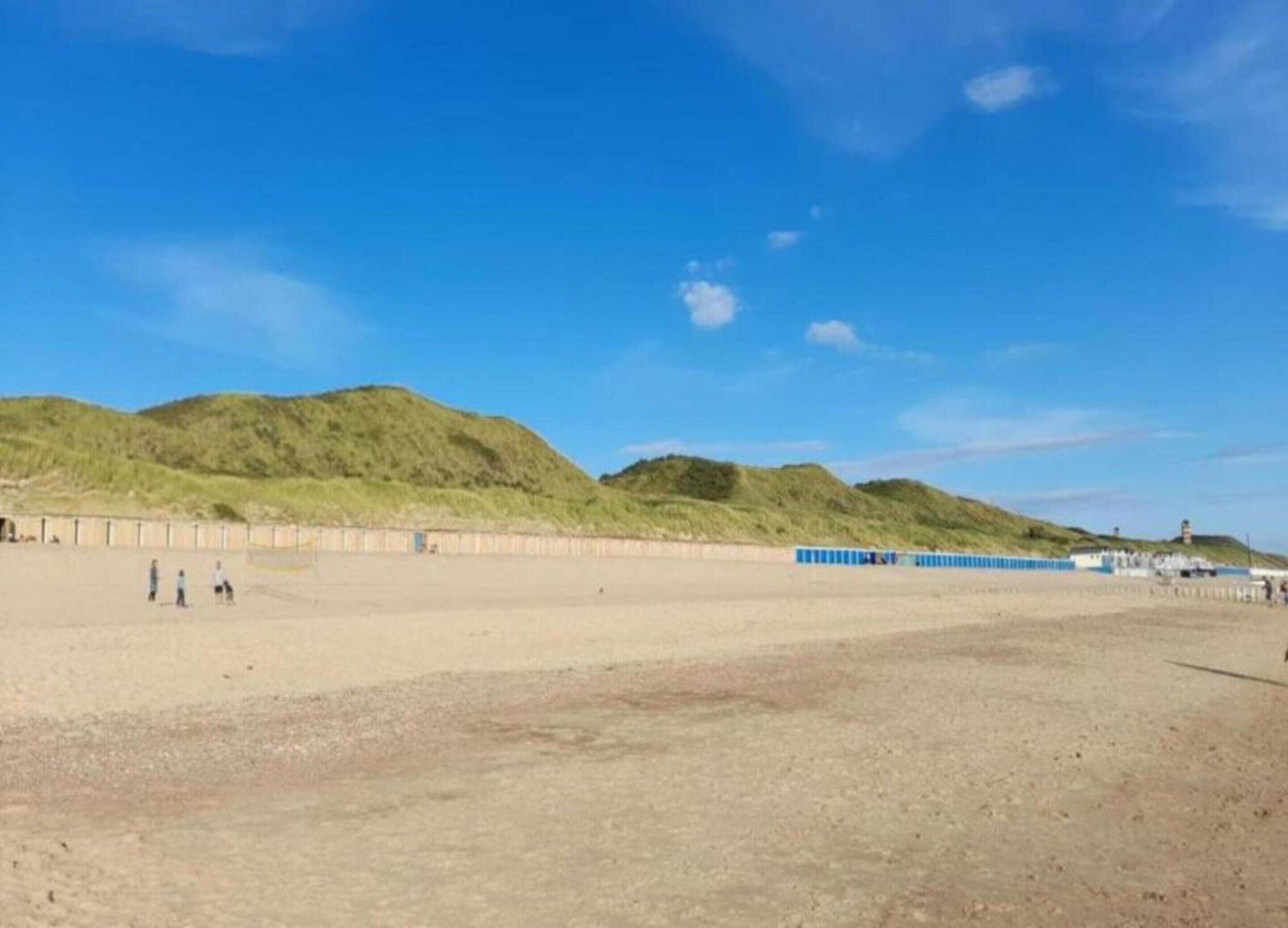 A sandy beach with a few people, a blue fence, and green hills in the background.