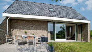 Une maison moderne avec une façade en briques, un toit en tuiles et un patio aménagé avec des meubles d’extérieur et des plantes.