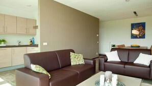 Un salon moderne avec deux canapés en cuir marron, une table basse agrémentée d’objets décoratifs et un coin cuisine avec des meubles de couleur claire.